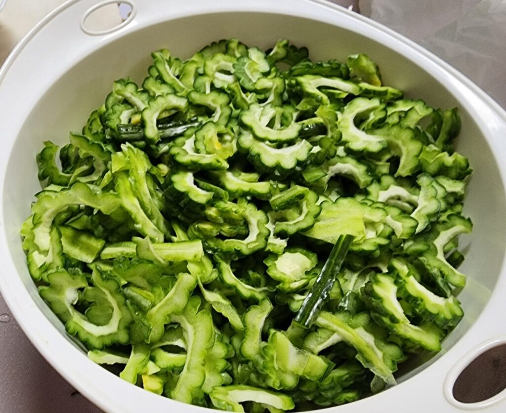 A close-up of a white bowl filled with raw, thinly sliced green bitter melon (ampalaya) with the seeds removed, ready for cooking.