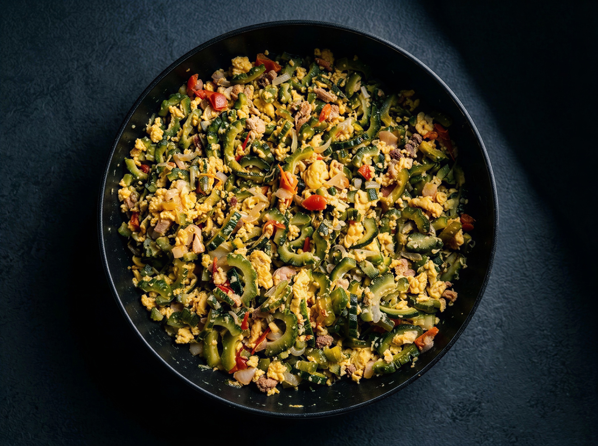 A black pan filled with freshly cooked Ampalaya con Itlog, featuring sliced green bitter melon, fluffy scrambled eggs, diced tomatoes, and onions on a dark textured background.